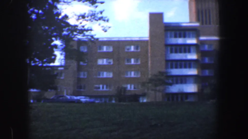 1960 Baltimore 8mm view of a brick apartment facade with rows of windows, trees, grass, and cars near the entrance area.