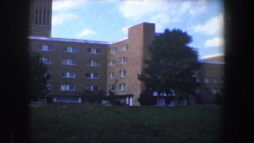 1960 Baltimore 8mm frame showing a brown brick building with a tall tower section, trees, lawn, and scattered clouds.