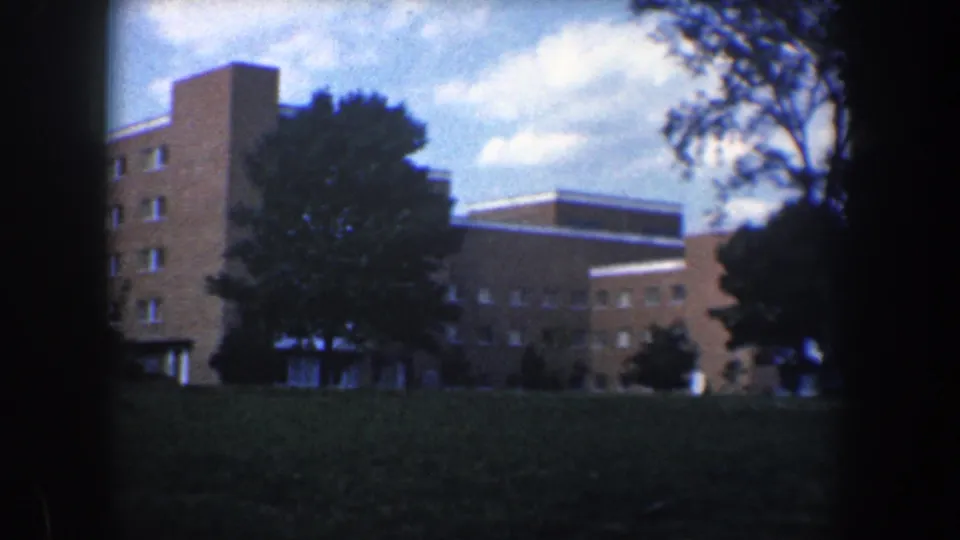 1960 Baltimore home-movie frame with the brick building centered behind trees and a wide grassy foreground.