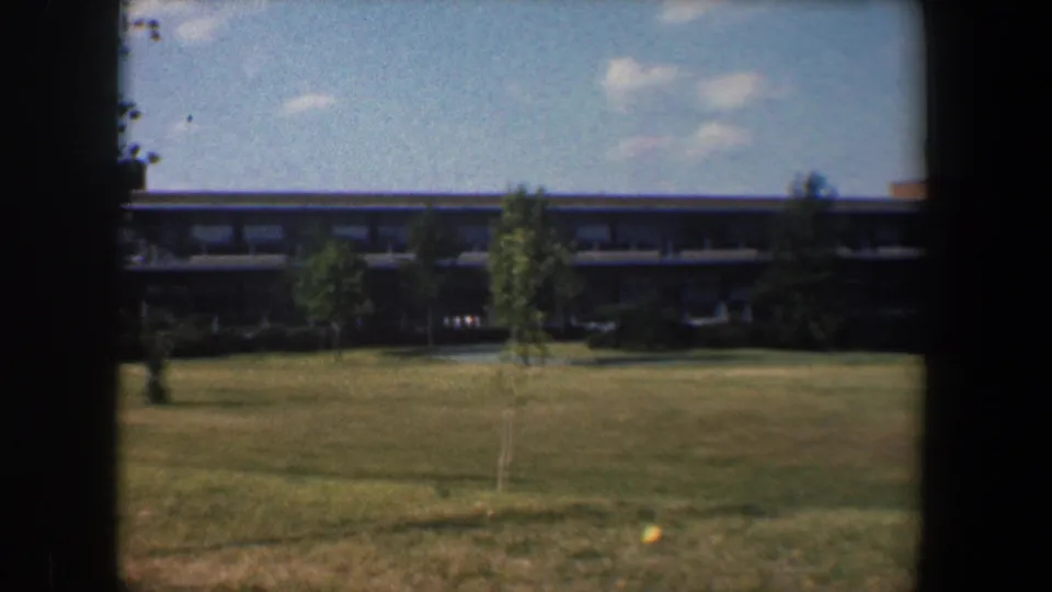 Baltimore campus exterior from 1960, showing lawn, young trees, a long low building and bright sky within 8mm frame borders.