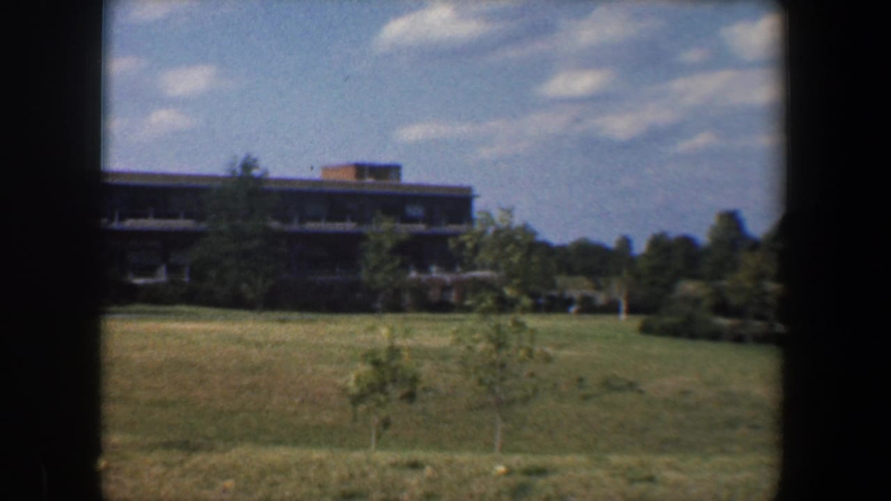 1960 Baltimore-area 8mm frame showing open grass, young trees, and a distant low multi-story building beyond the field.