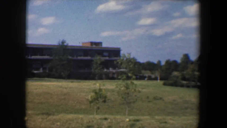 1960 Baltimore-area 8mm frame showing open grass, young trees, and a distant low multi-story building beyond the field.