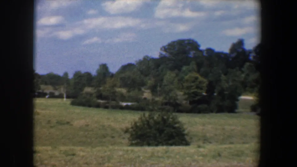 1960 Baltimore-area frame showing a sloped grassy field with shrubs, mature trees, and a possible road or path beyond the open ground.
