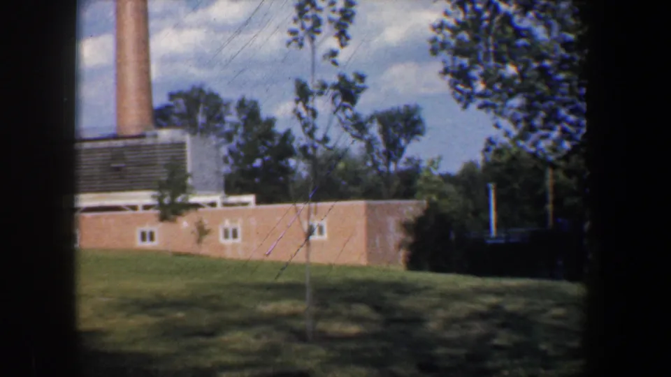 1960 Baltimore-area 8mm frame showing a brick industrial structure, tall smokestack, utility lines, grass, and surrounding trees.