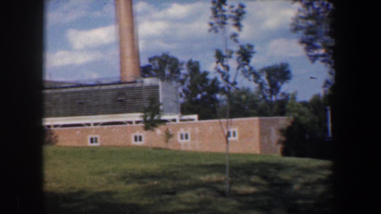 1960 Baltimore 8mm frame showing a brick industrial-style building, tall smokestack, lawn, young trees, and blue sky.