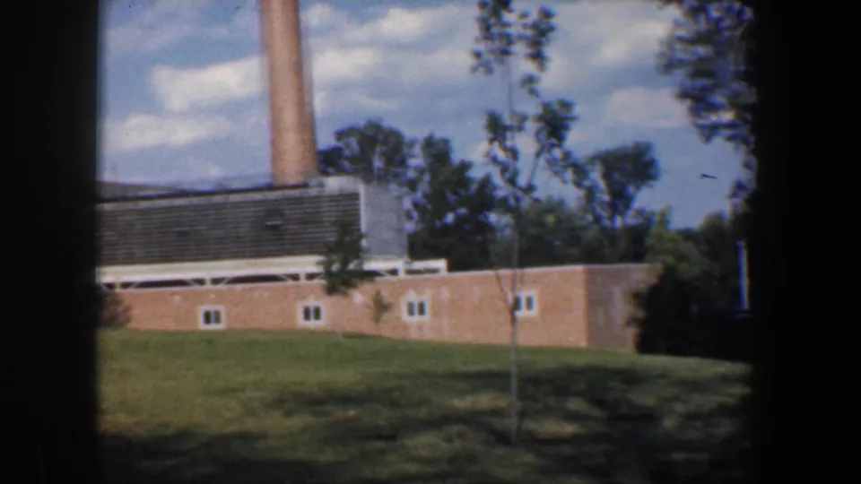 1960 Baltimore 8mm frame showing a brick industrial-style building, tall smokestack, lawn, young trees, and blue sky.