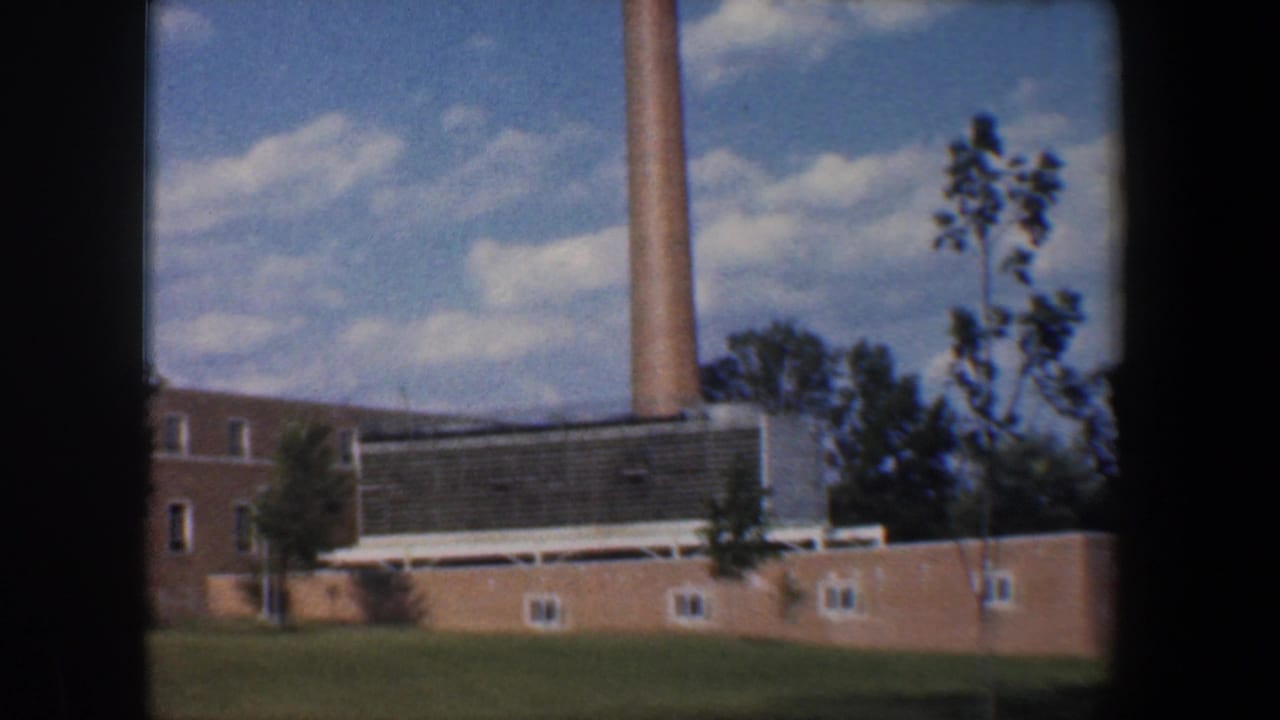 Tall smokestack rises behind a brick building with trees and a cloudy blue sky.