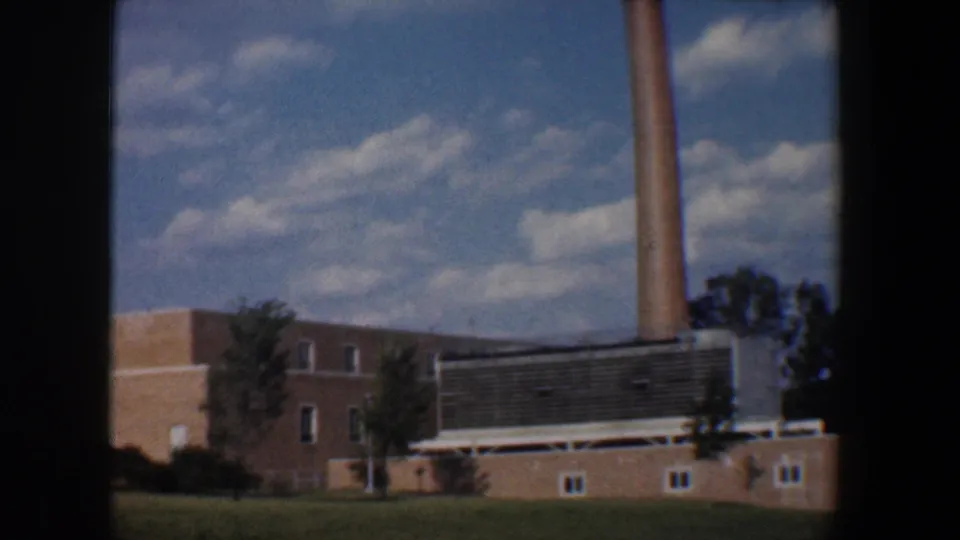 1960 Baltimore 8mm frame of a brick industrial-looking structure with smokestack, louvered section, lawn, and clouded sky.
