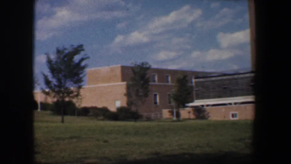 1960 Baltimore 8mm wide view of a brick building, landscaped grass, small trees, dark louvered panel, and cloudy blue sky.