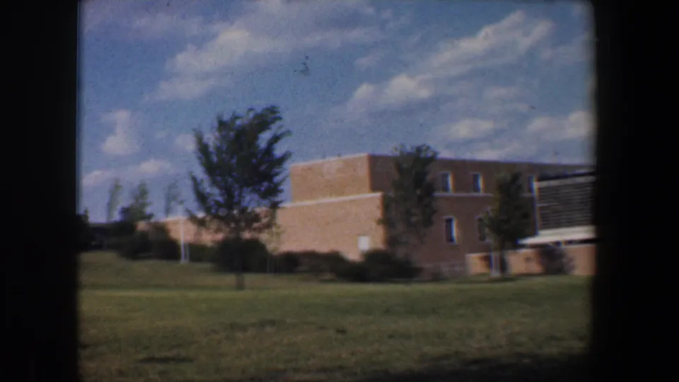1960 Baltimore-area 8mm frame showing a tan brick institutional-looking building, lawn, trees, and a bright sky.