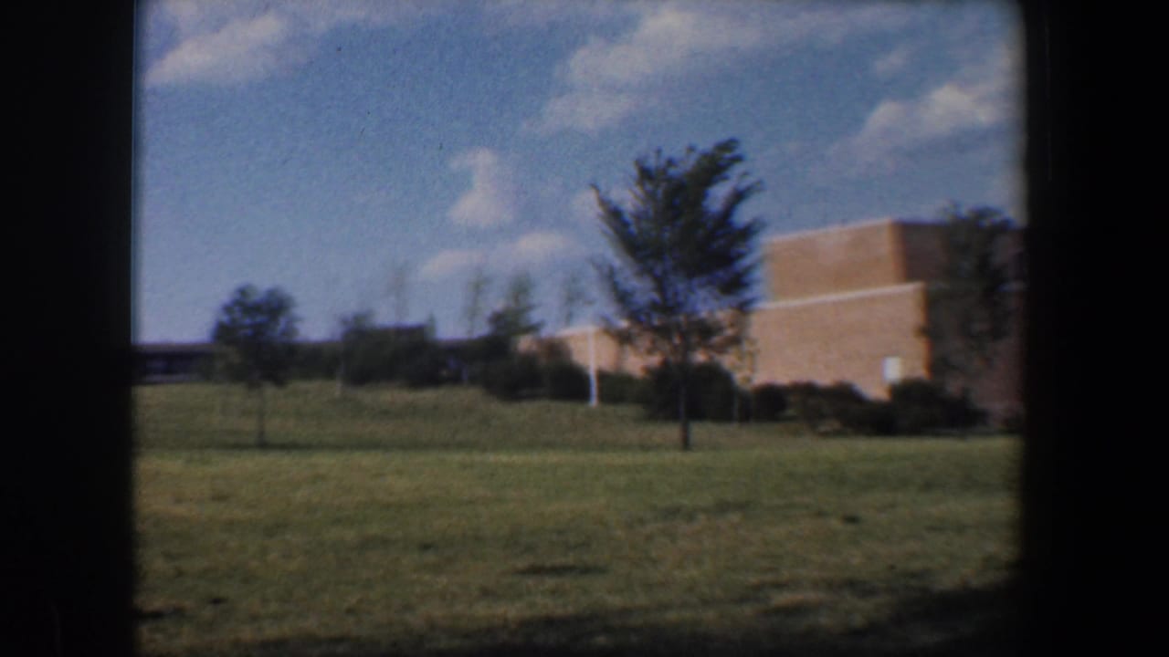 Open lawn with a tan brick building at right and trees against blue sky.