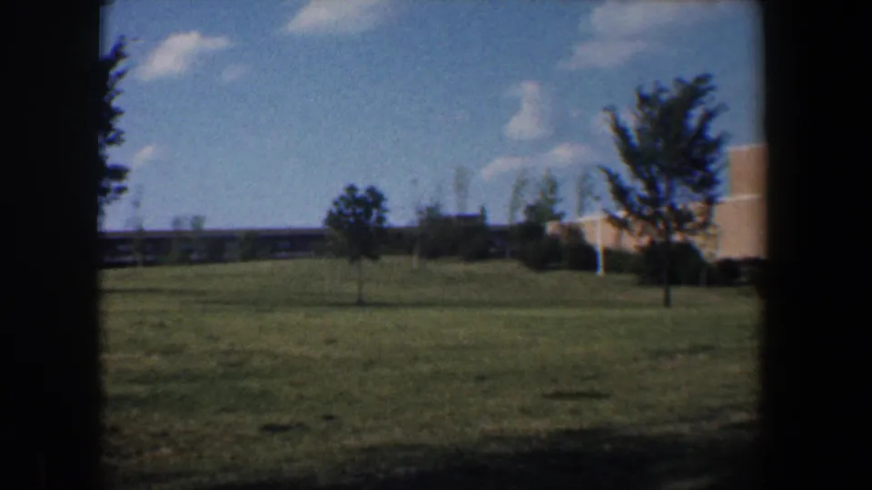 1960 Baltimore-area 8mm view across landscaped grounds toward trees, shrubs, and a partial tan brick building.