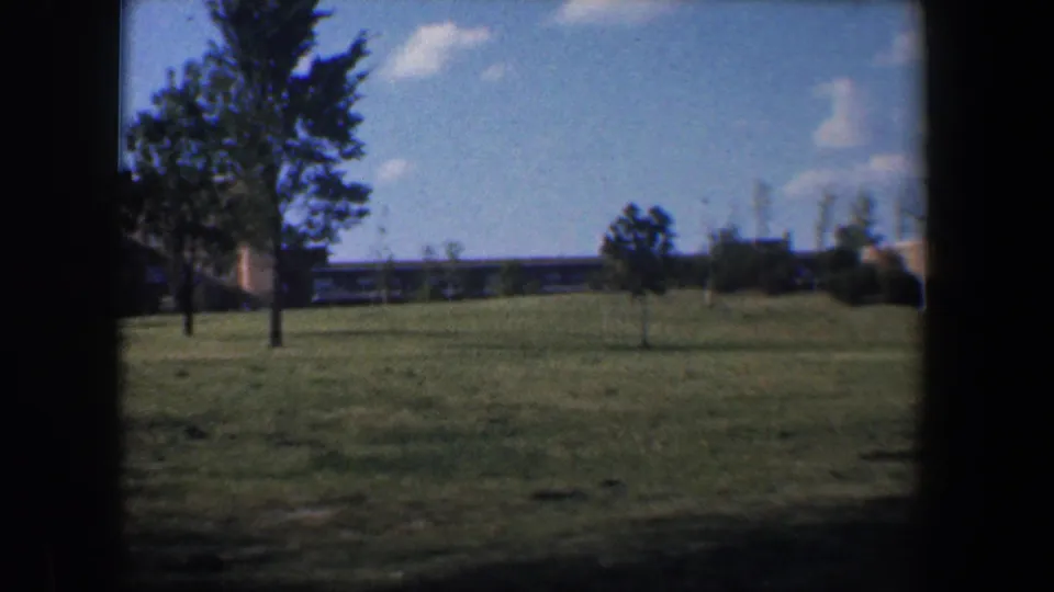 1960 Baltimore-area frame with lawn, several trees, distant low structures, and a bright blue sky on 8mm film.