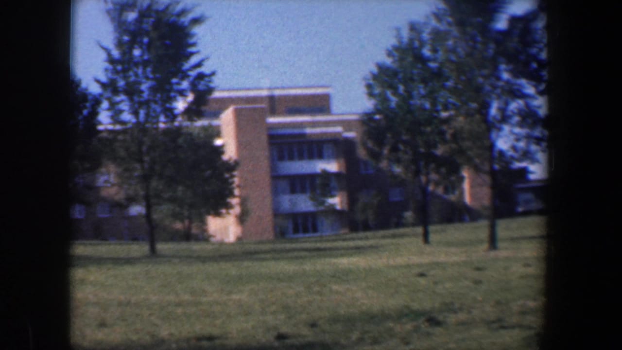 Brick building wing with window bands viewed across open grass and trees.