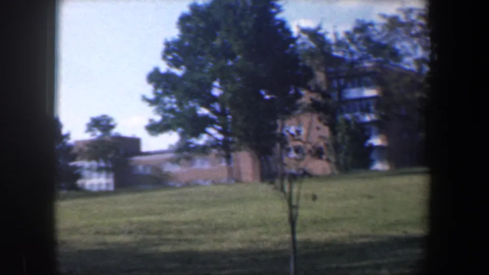 1960 Baltimore 8mm frame with a small tree in the foreground, larger trees, lawn, and brick building facades.