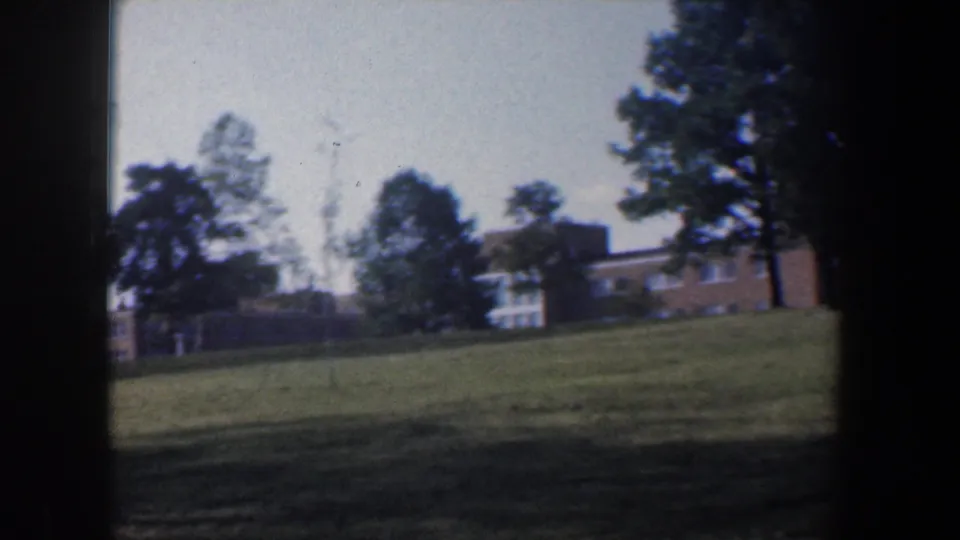 Baltimore 1960 home-movie view of a grassy slope, trees, and distant brick buildings on institutional grounds.