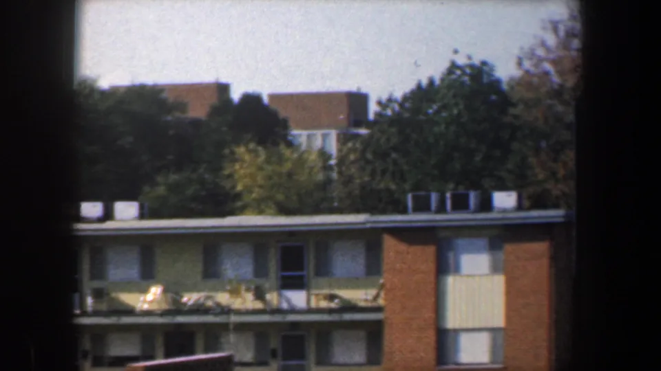 1960 Baltimore frame showing a low building with rooftop units below taller tan brick structures and surrounding trees.