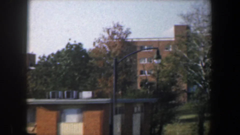 1960 Baltimore exterior with a streetlamp, low roofline, trees, and a taller tan brick building rising behind grass.