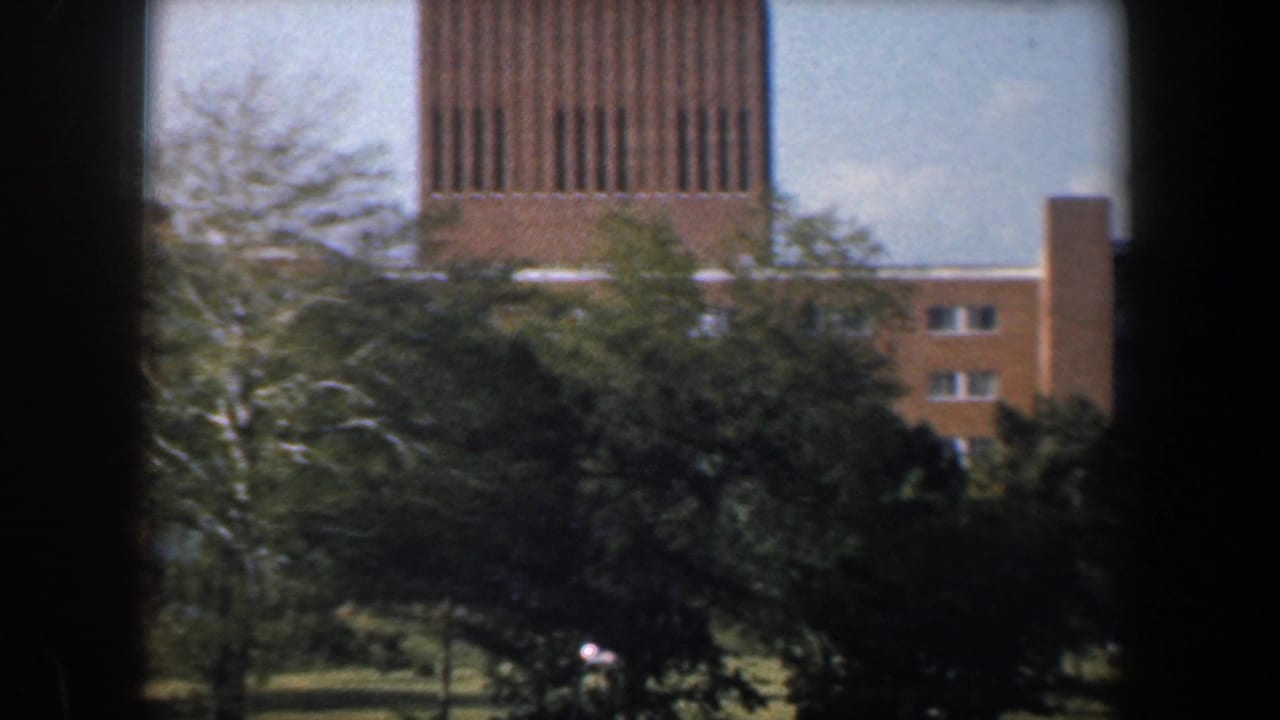 Brick apartment block with a tall slatted tower partially obscured by trees and dark vertical framing.