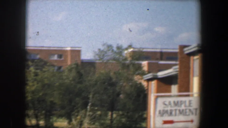 1960 Baltimore frame with a sample apartment sign beside trees and tan brick residential buildings under a clear sky.