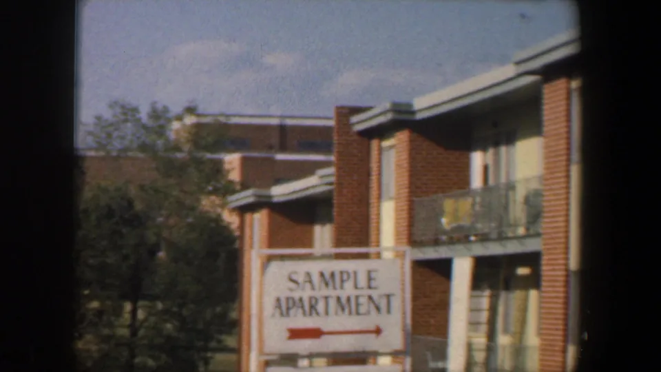 1960 Baltimore home-movie frame of a brick apartment complex and white SAMPLE APARTMENT sign viewed through dark window bars.