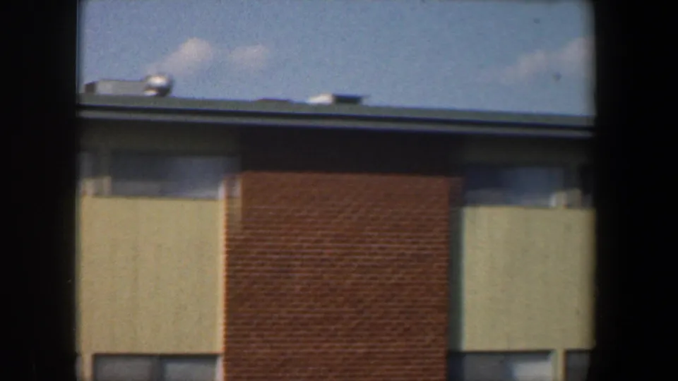 1960 Baltimore frame showing a sunlit apartment facade with red brick, pale green panels, and small upper windows.