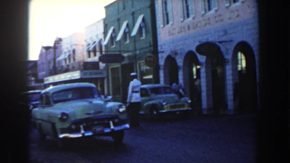 Period cars move along a Nassau street as a white-uniformed officer stands near arched shopfronts.