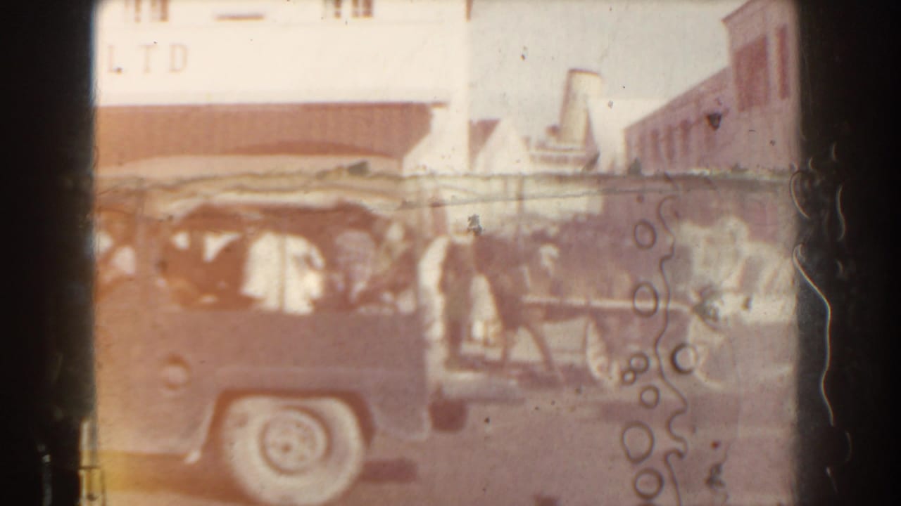 Vintage car passes in front of a garage storefront with a carriage partly visible behind it.