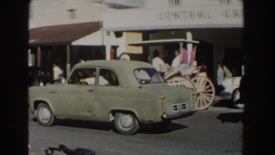 Green mid-century car moves past a carriage and pedestrians outside Central Garage Ltd in Nassau, Bahamas.
