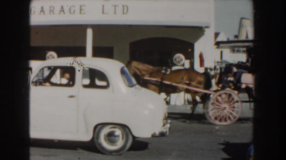White vintage car in the foreground with a horse-drawn carriage and Central Garage Ltd facade behind it.