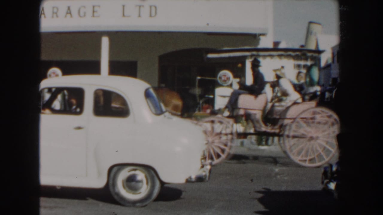 Horse-drawn carriage beside a white car outside a garage building.