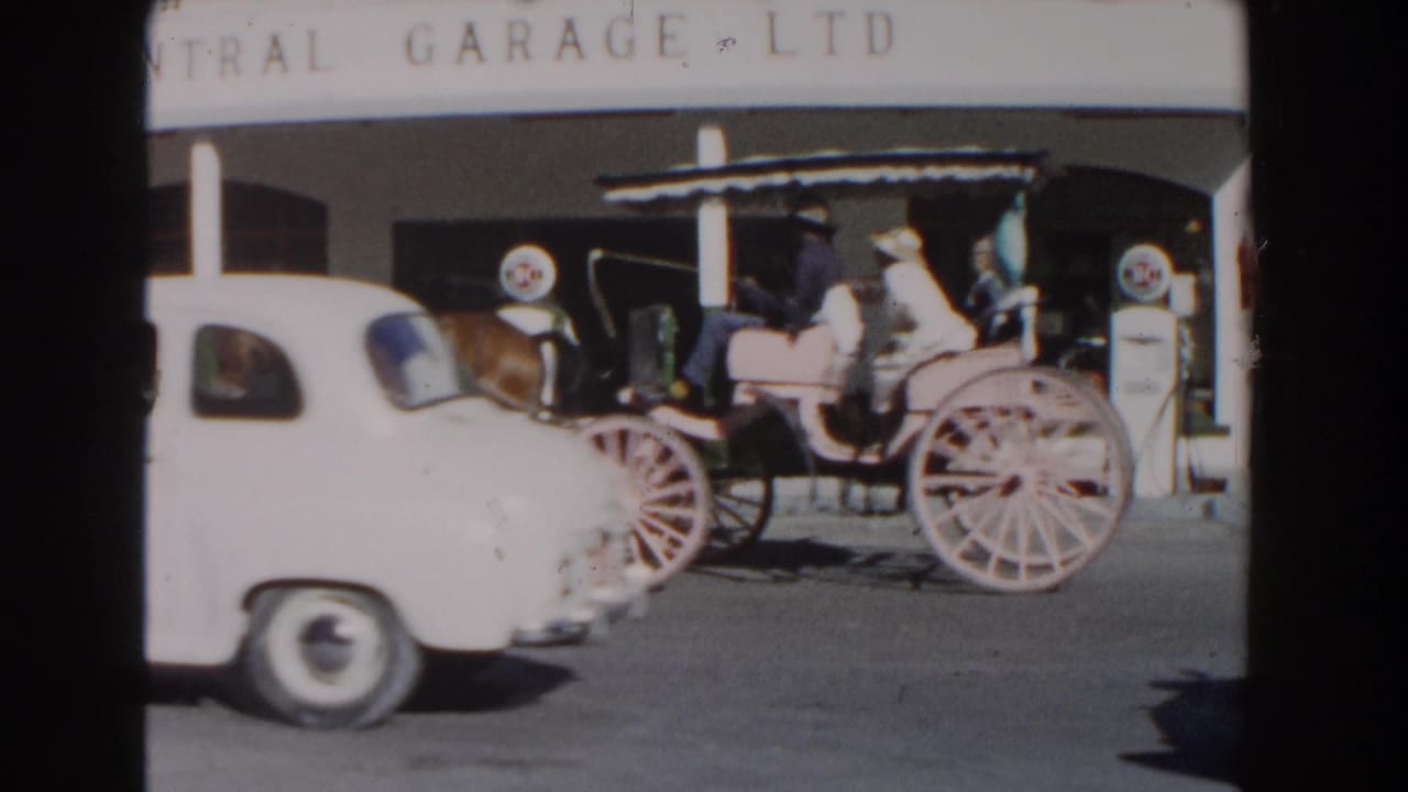 Horse carriage with passengers passing fuel pumps and a white car.
