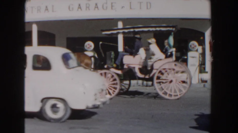 A horse-drawn carriage carrying passengers passes fuel pumps and a white mid-century car on a Nassau street in the 1961 home-movie clip.