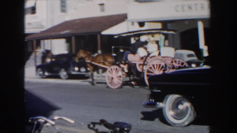 The carriage continues along a sunny Nassau street while a dark mid-century car occupies the foreground and the garage building remains visible behind it.