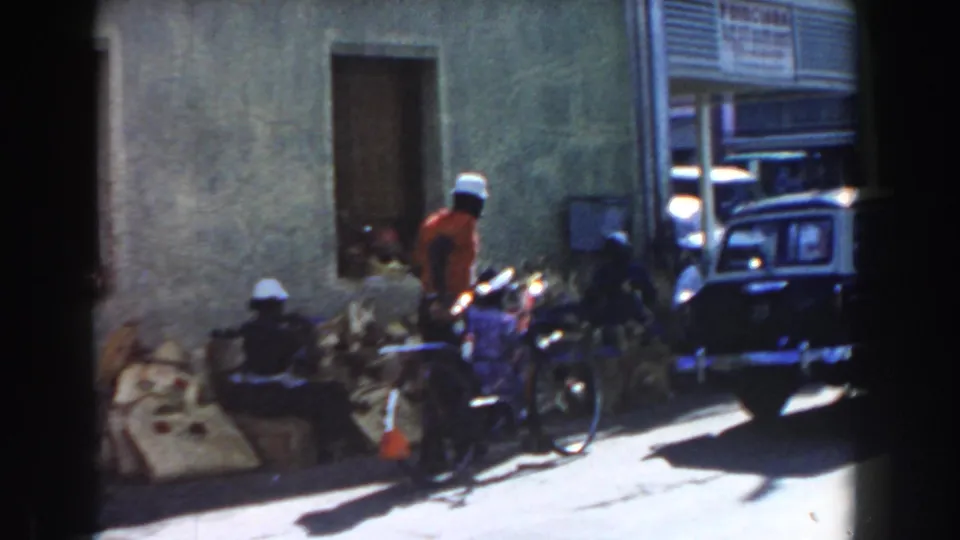 Nassau street scene with pedestrians, bicycles, stacked sacks and a dark vintage car beside a building.