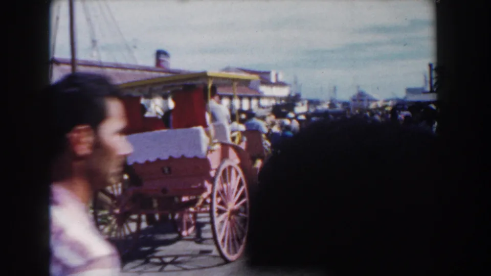 Nassau home-movie frame showing a red carriage, a blurred passerby and a crowded street beyond.
