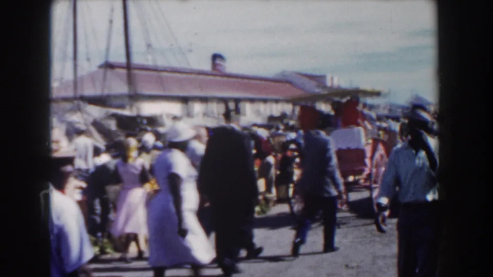 1961 Nassau street activity with pedestrians, a red horse-drawn carriage, summer clothing, and harbor-side structures in handheld 8mm footage.