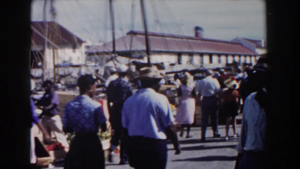 Nassau street in 1961 with pedestrians walking past a carriage wheel, nearby masts, and low waterfront-style buildings in grainy 8mm color.