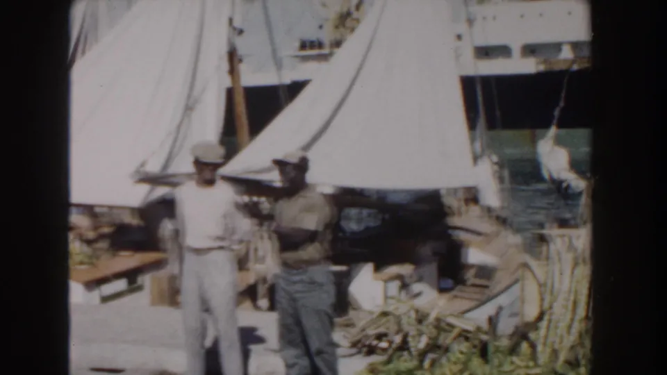 1961 Nassau harbor scene with men in workwear near large white canvas shade tents, barrels, ropes, and sailboats.