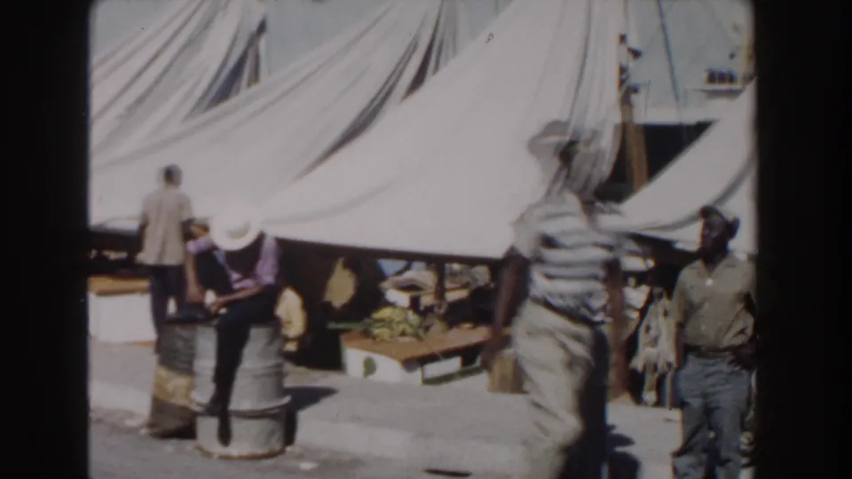 Men at a Nassau waterfront work area stand and sit around barrels and tables under large white canvas tents.