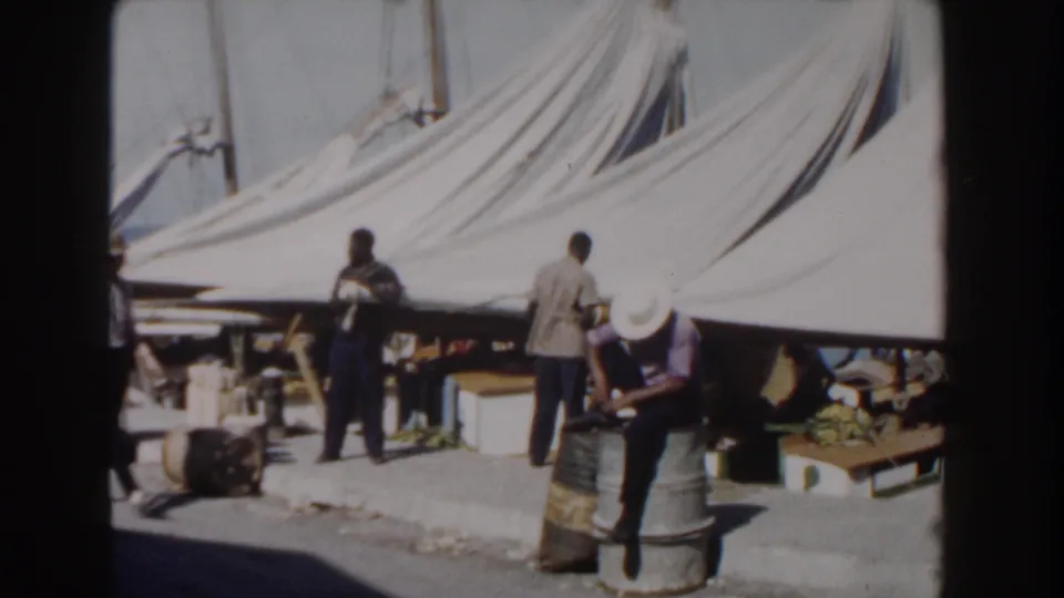 Candid 8mm view of men in Nassau near canvas shade, barrels, boxes, and boat masts in the harbor background.