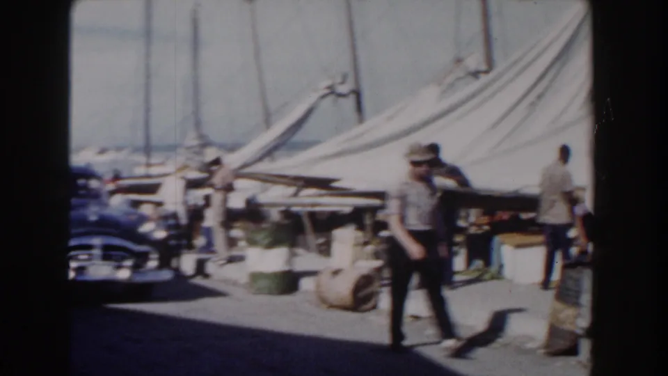 Nassau harbor frame with a man walking in front of canvas tents, barrels, a blue car, and sailboat masts.