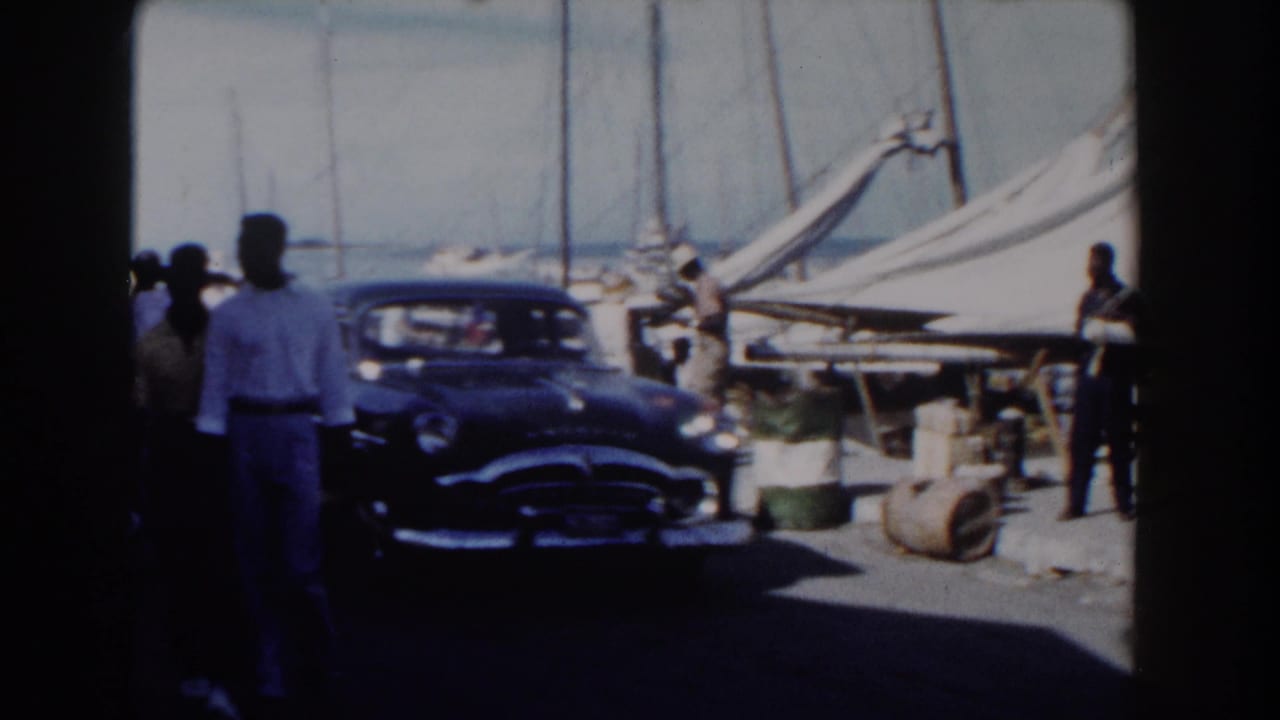 1961 Nassau pier scene with adults walking near a dark classic car, dockside barrels, and sailboats in bright daylight.