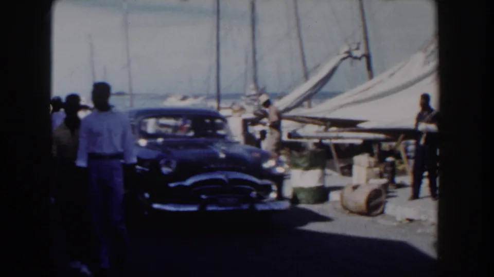 1961 Nassau pier scene with adults walking near a dark classic car, dockside barrels, and sailboats in bright daylight.