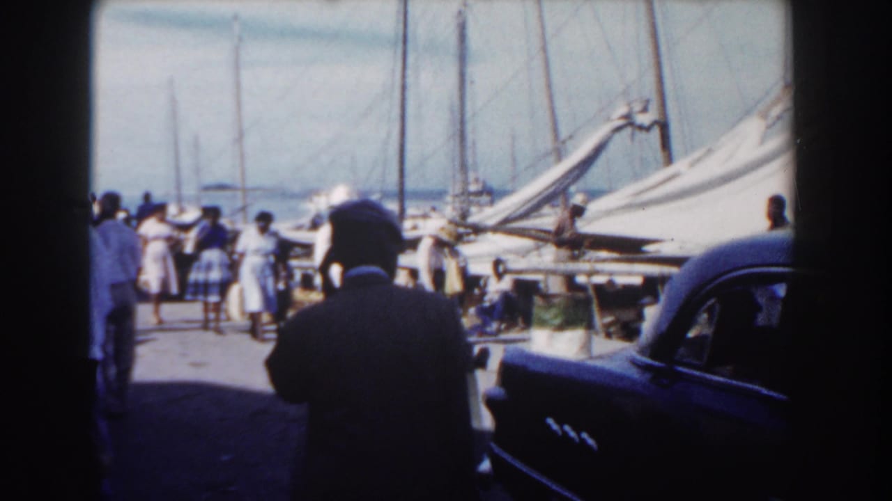 Crowd on a pier with sailboats and part of a dark car at the edge.