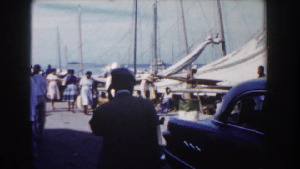 Adults gather and walk on a Nassau pier in 1961, with sailboat masts, docked boats, and a dark car partly visible.