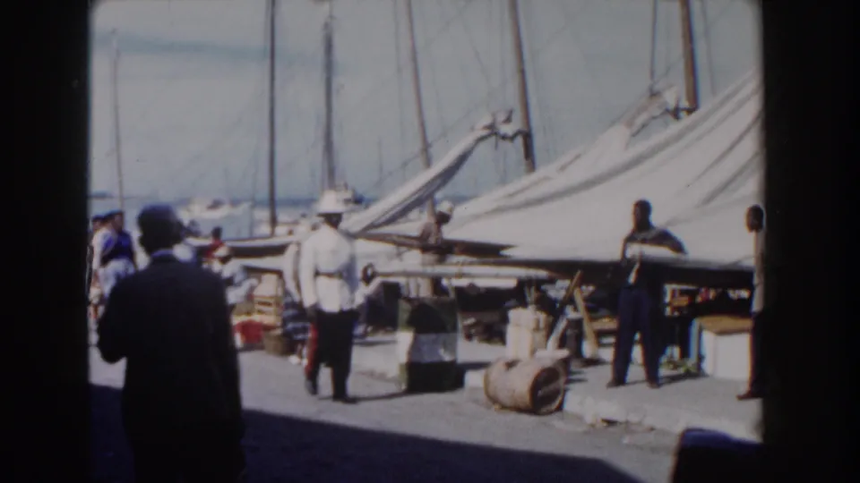 1961 Nassau dock view with adults in light clothing, sailboats, barrels, and merchandise-like items along the pier.