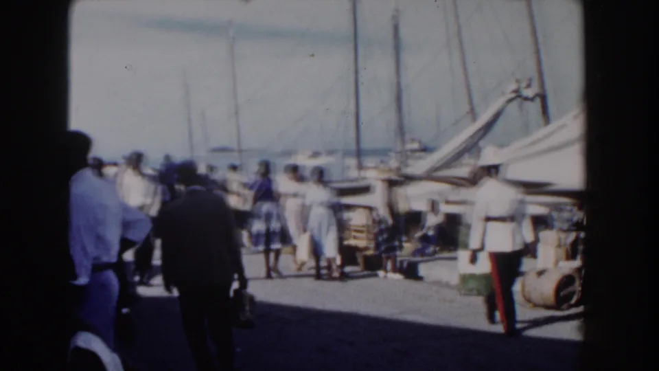 Nassau Bahamas pier activity in 1961, showing adults near sailboats, barrels, dock materials, and the harbor beyond.
