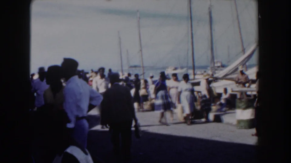 1961 Nassau harbor scene with people waiting near a pier, shaded foreground figures, and sailboat masts beyond.