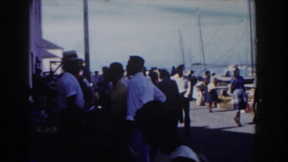 Nassau dockside crowd seen from shade, with men and women near moored sailboats under bright harbor light.